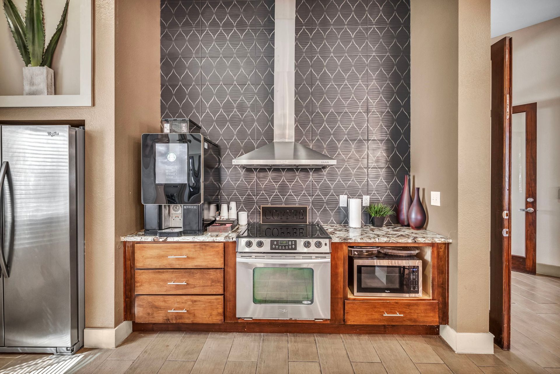 Kitchenette with espresso machine, stove, microwave, and refrigerator. Dark tile backsplash, wood cabinets, and stainless steel appliances.