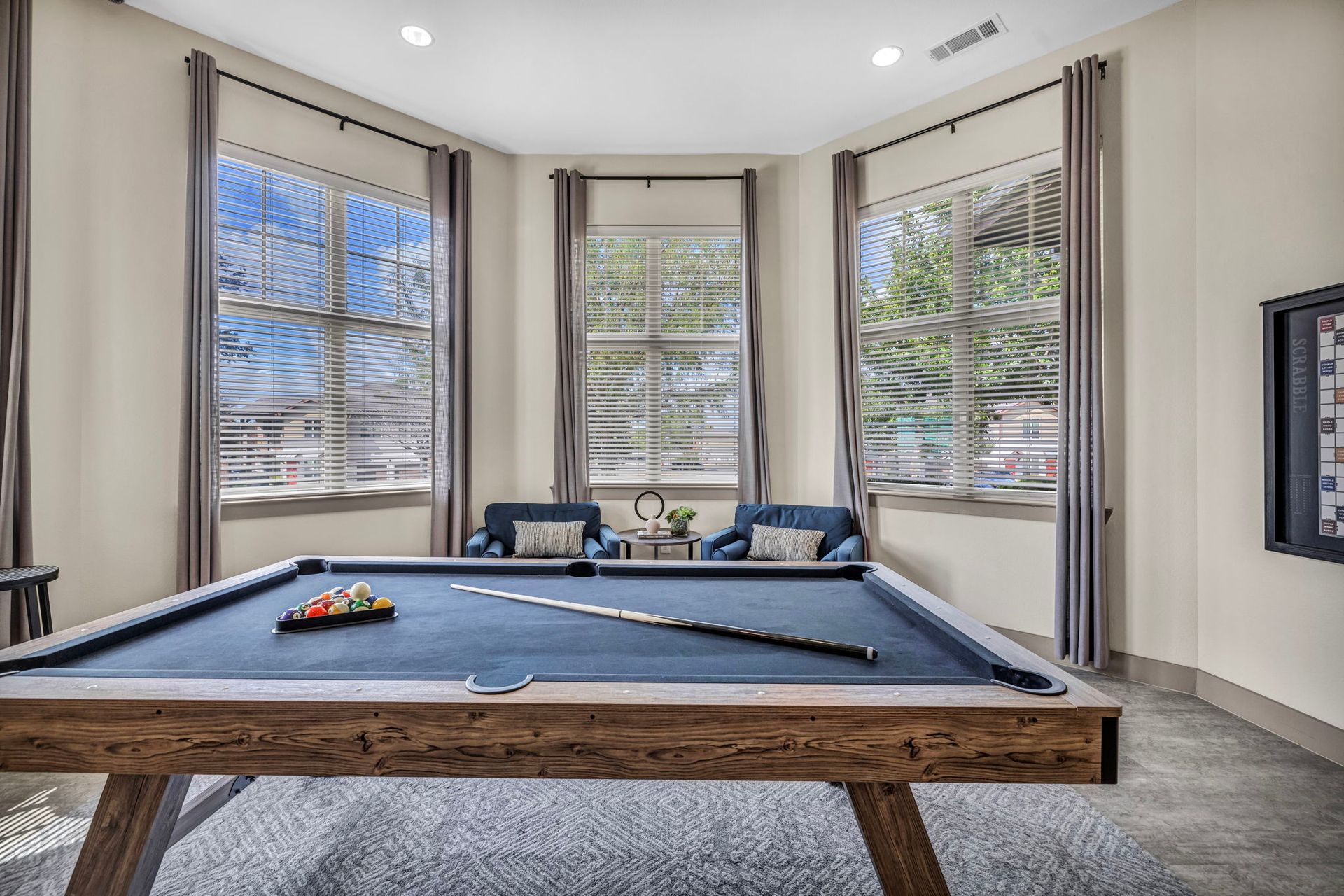 Pool table in a room with three large windows, two blue chairs, and gray curtains.