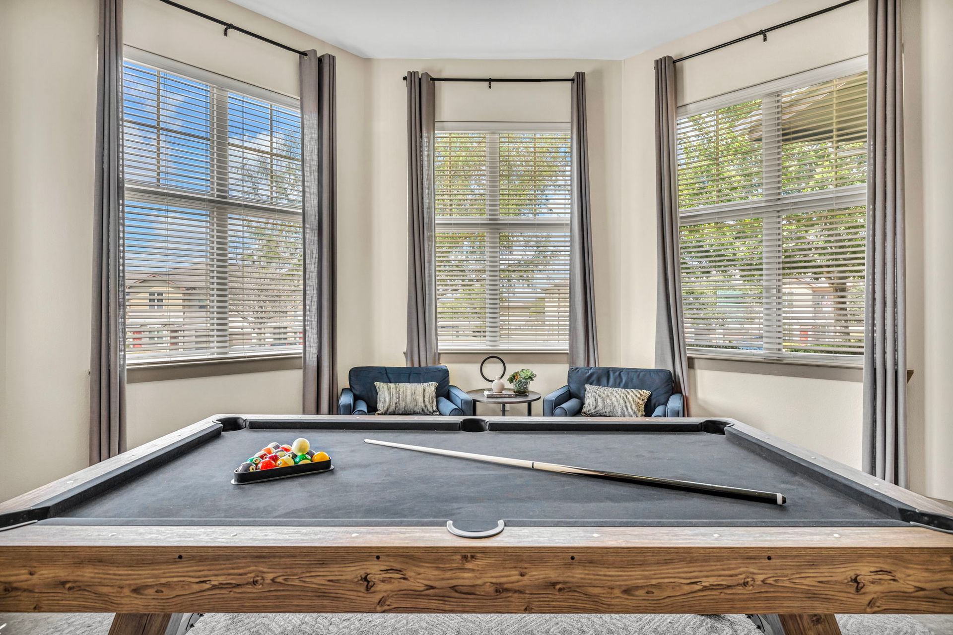 Pool table in a room with large windows, two armchairs, and pool cues, inviting a game.