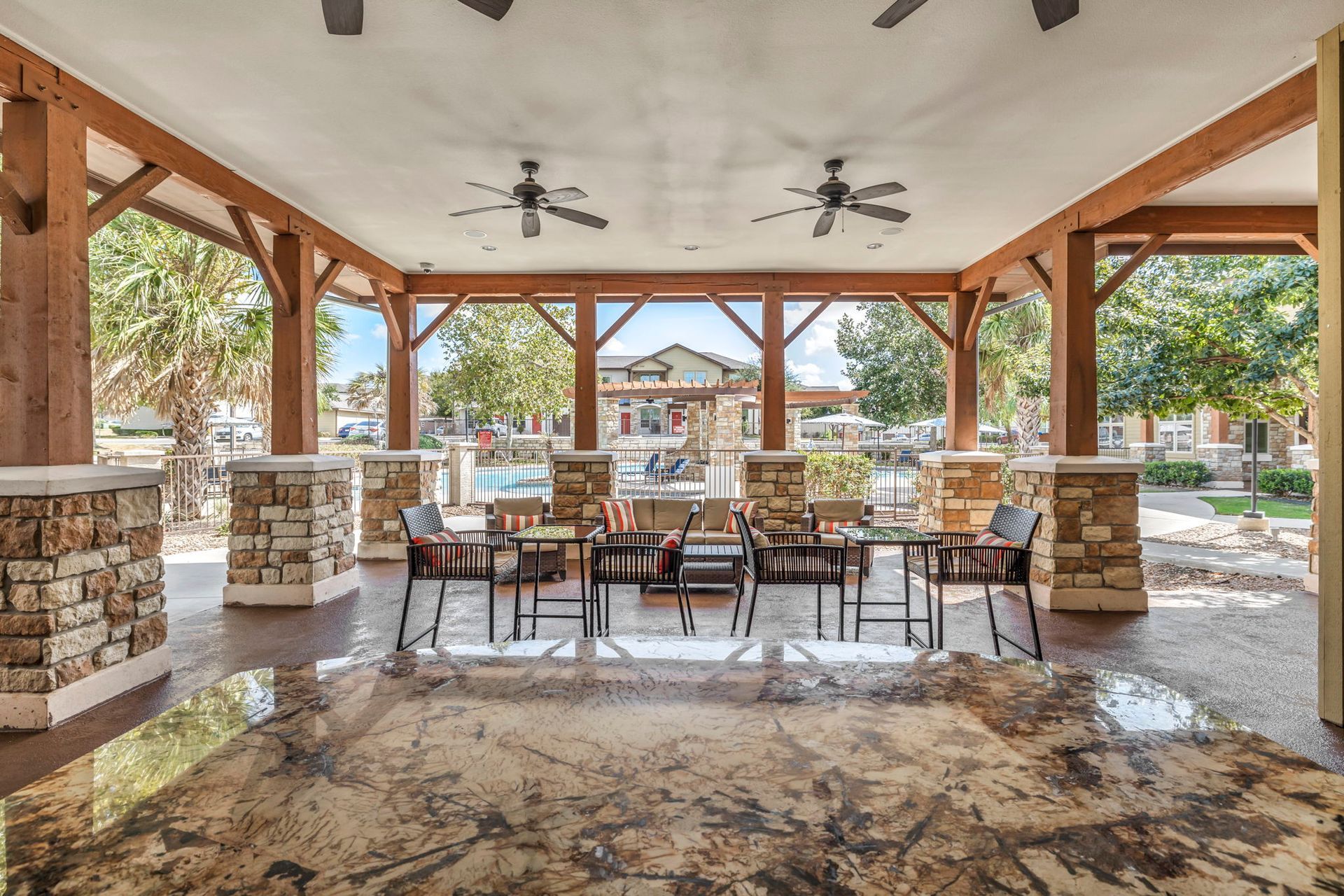 Covered outdoor seating area with tables and chairs near a pool. Brown wood beams and stone columns.