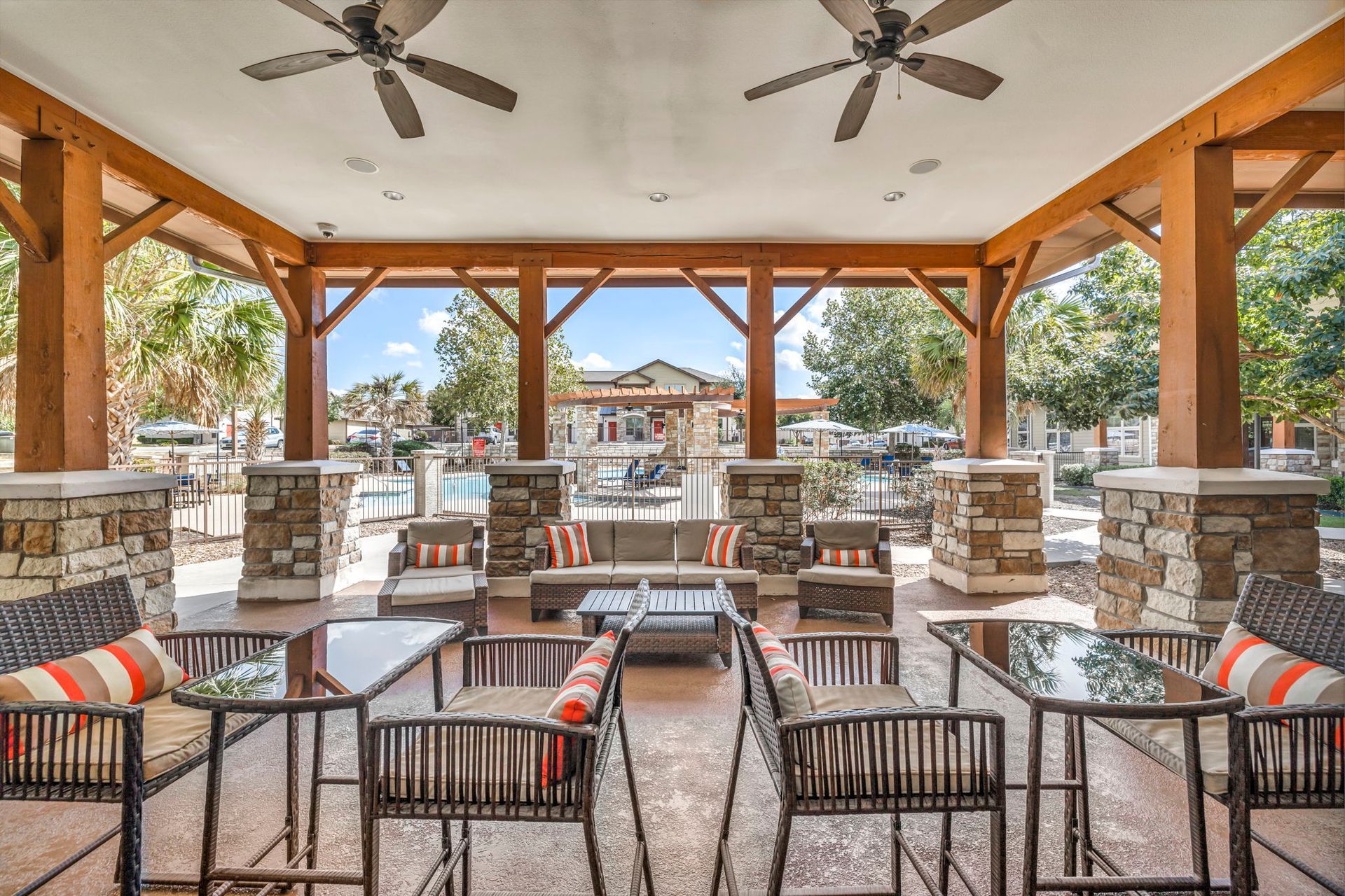 Outdoor covered patio with seating and ceiling fans. Brown wood beams, stone columns, and neutral furniture.