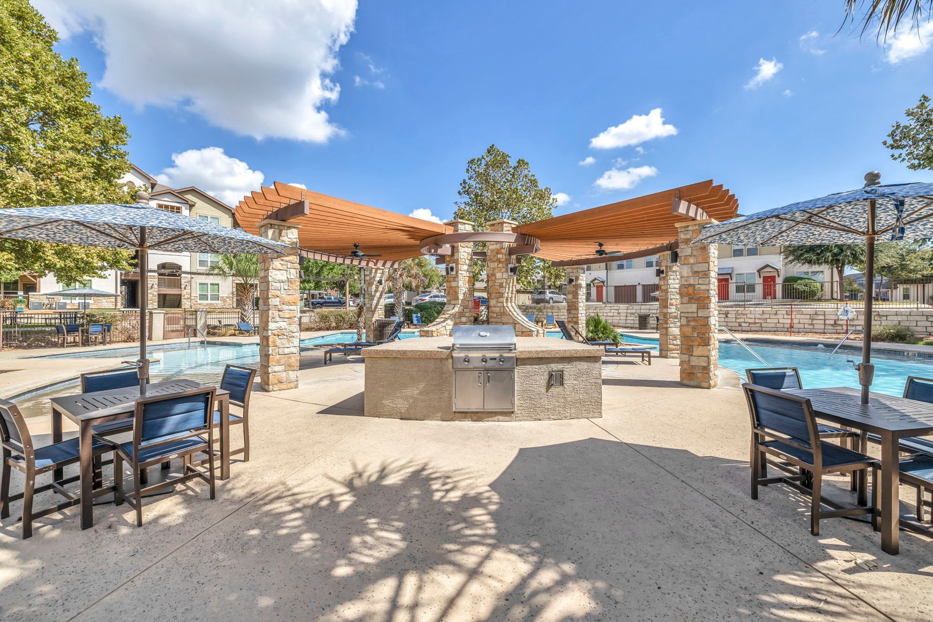 Outdoor kitchen area with pool, tables, and umbrellas under a sunny sky.