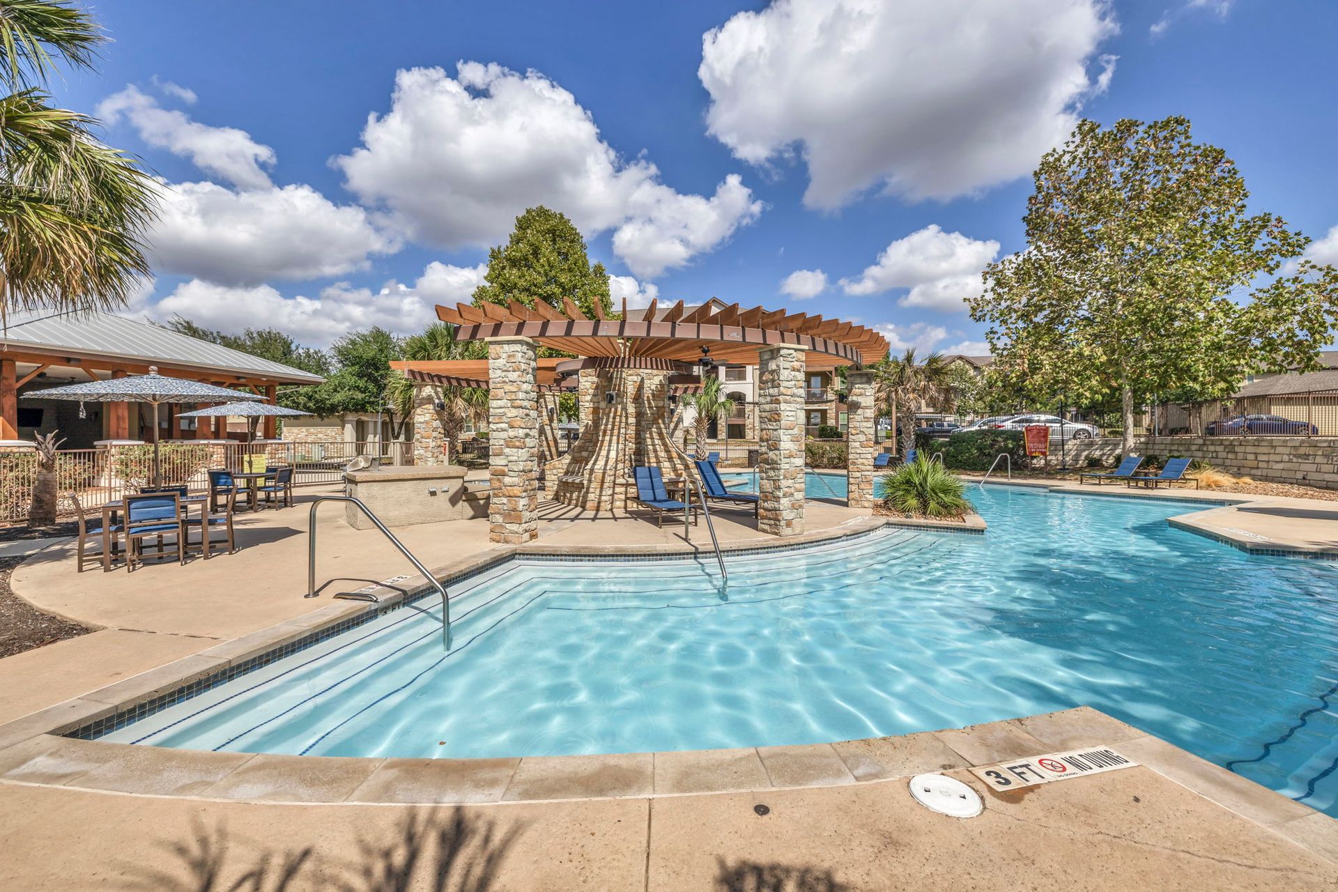 Pool area with blue water, stone pergola, and lounge chairs under a sunny sky.