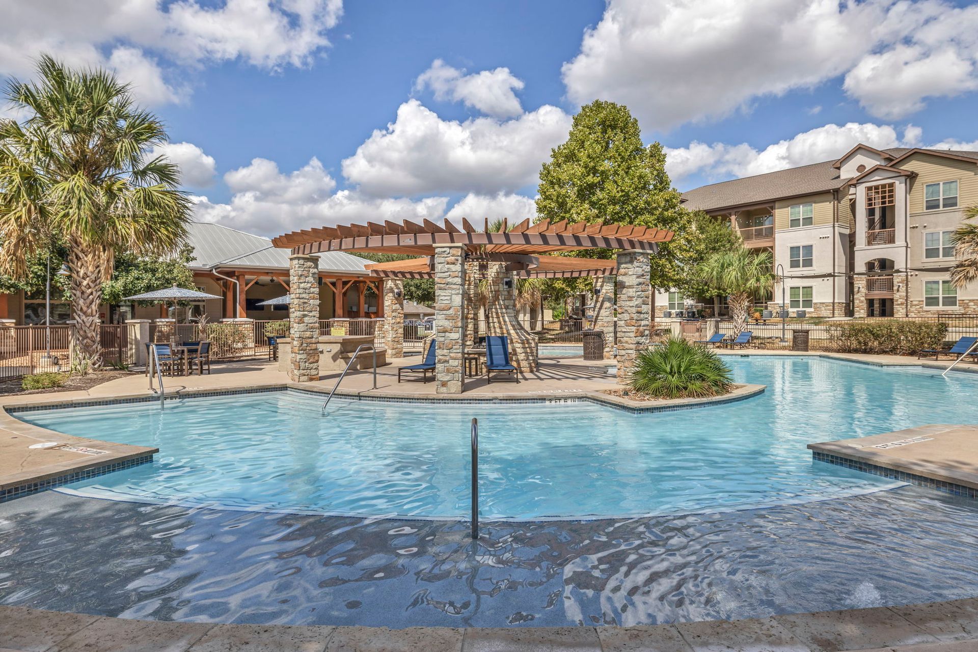 Swimming pool with stone pergola and apartment building, sunny day.