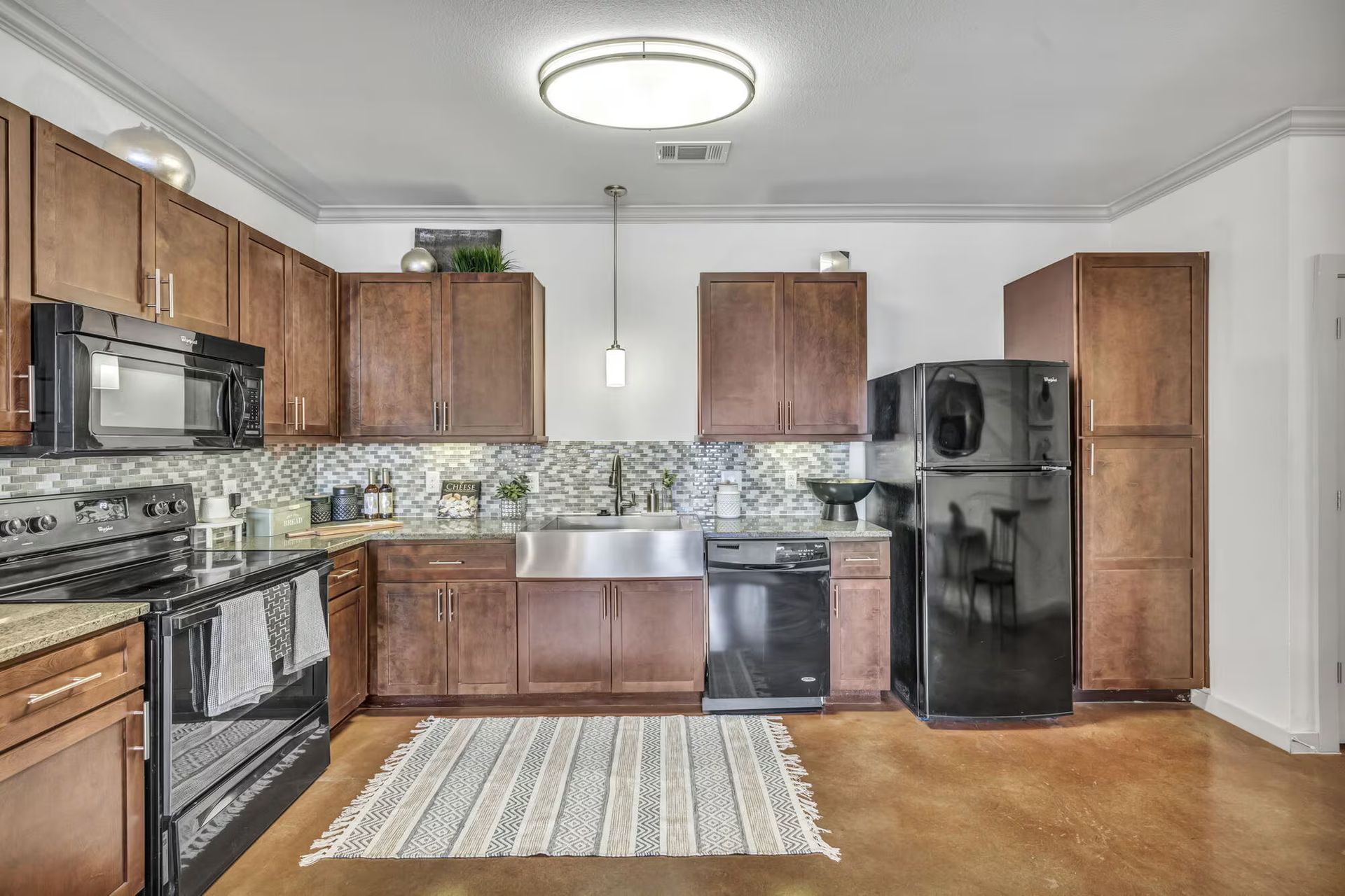 A modern kitchen with dark wood cabinets, stainless steel sink, black appliances, and a patterned rug.