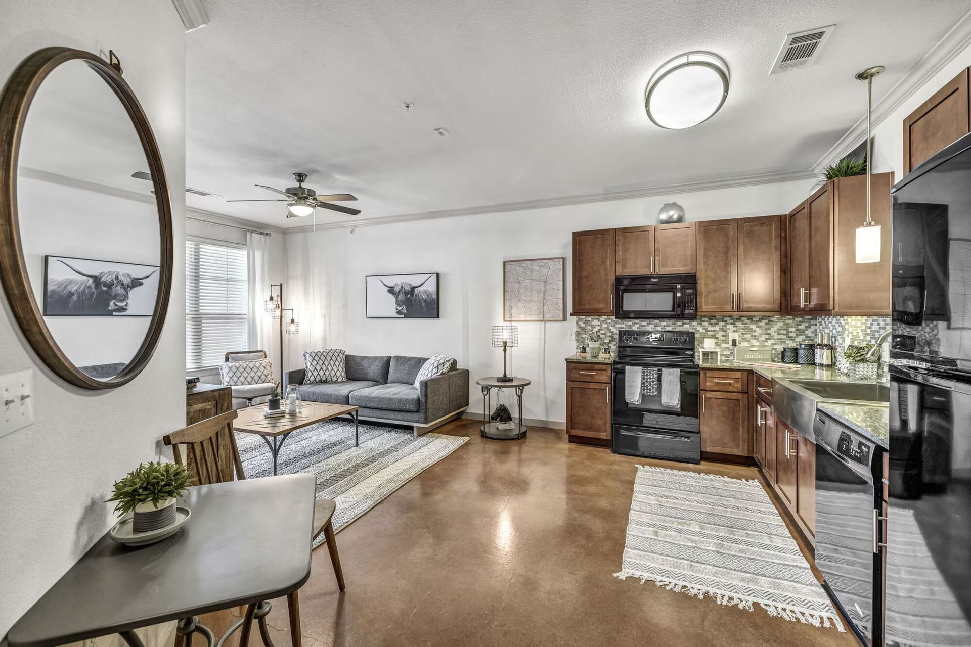 Open-concept living space: kitchen with brown cabinets, living room with grey sofa, and dining area with a round mirror.