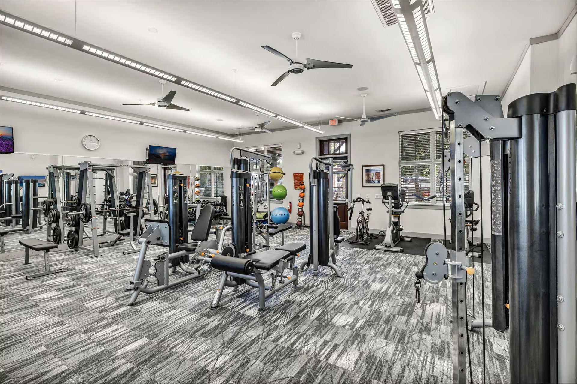 A fitness center with various exercise machines, ceiling fans, and gray-patterned carpet.