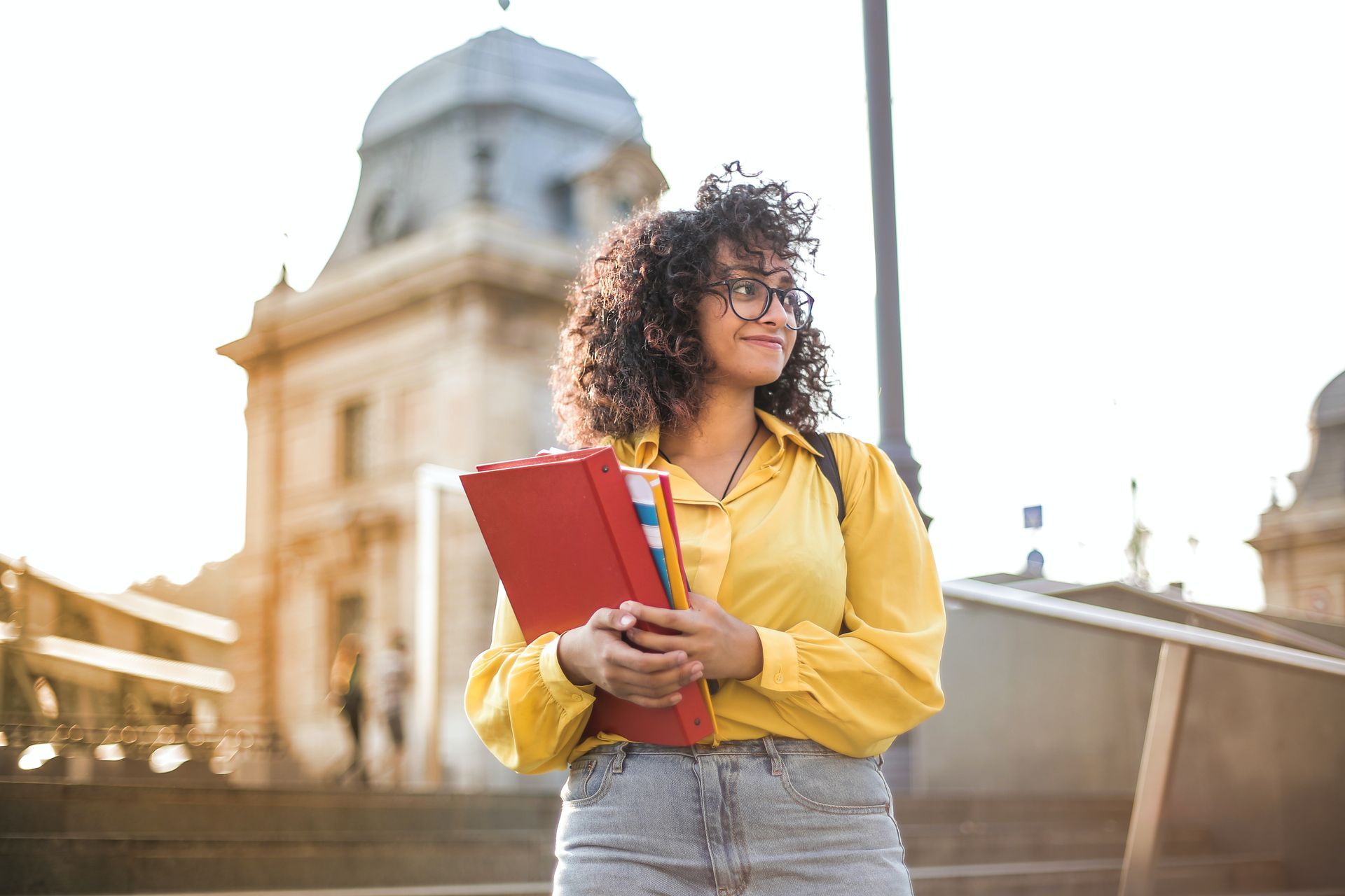 Young woman with curly hair, yellow shirt, holding red books, smiling outside a building.