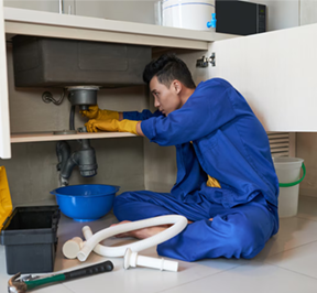 Plumber in blue overalls under a kitchen sink, wearing gloves, working on pipes.