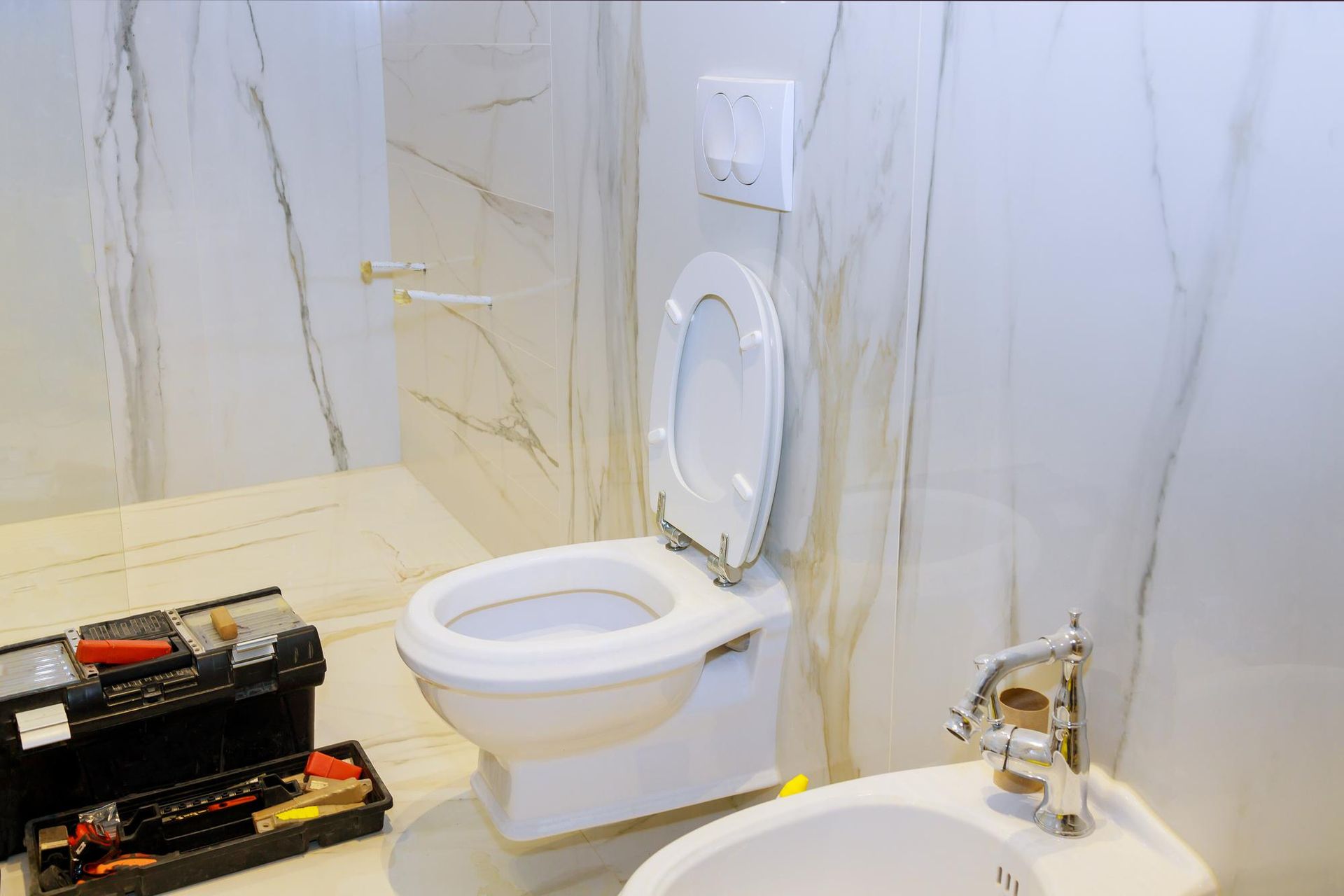 Toilet being repaired in bathroom with a toolbox, bidet, and marble-like wall.