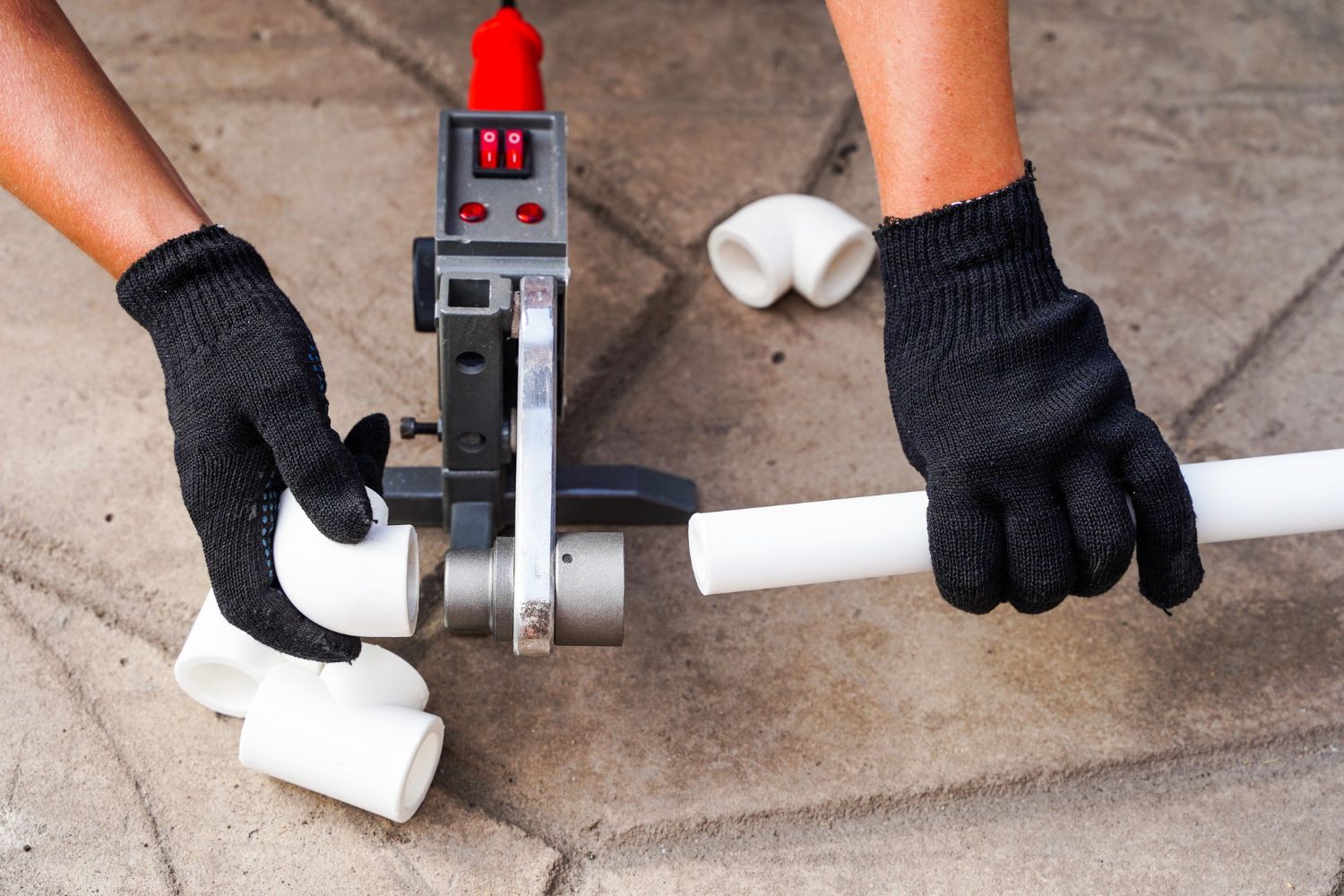 Hands using a pipe fusion tool to connect white PVC pipes and fittings on a stone surface.