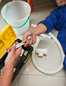 Plumber receiving a tool from someone while working under a sink. Tools, bucket, and pipes visible.