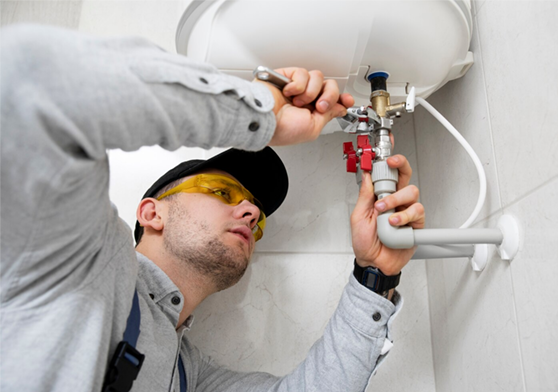 Plumber wearing safety glasses works on plumbing under a white appliance, with a grey shirt and black cap.
