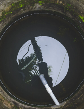 Looking down into a concrete well filled with water, reflecting the sky and nearby trees. A white pipe extends into the water.