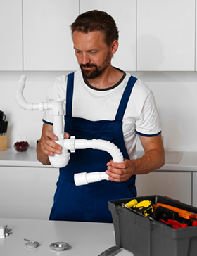 Plumber in blue overalls examining sink drain pipes in a white kitchen, tool box on the counter.