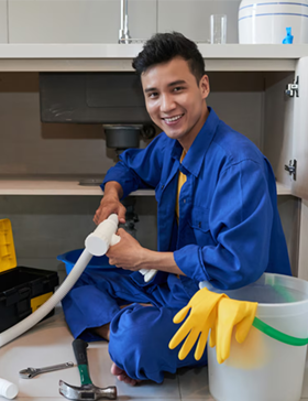 Plumber in blue overalls smiling while working under a kitchen sink. Yellow gloves and tools nearby.