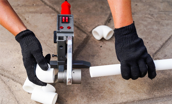 Hands in black gloves using a PVC pipe welding tool. Elbow fittings and pipe on concrete.