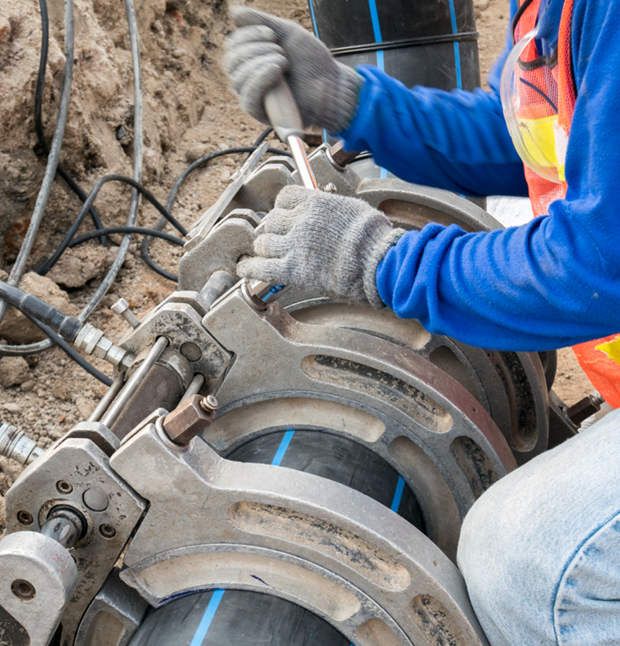 Person in work clothes using a tool to join black pipes, outdoors.