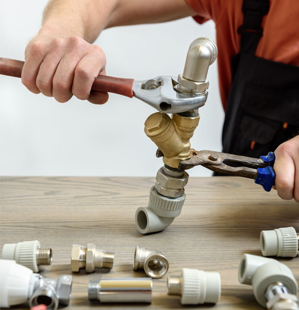 Plumber using wrenches to assemble plumbing fittings on a wooden surface, with various parts nearby.