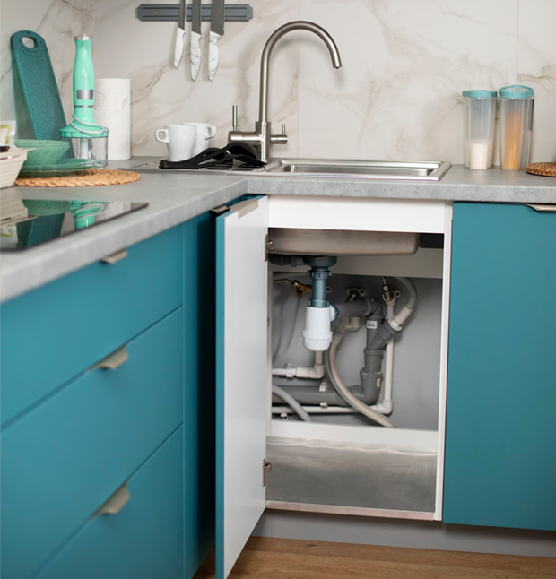 Kitchen sink cabinet open, revealing plumbing. Turquoise cabinets, stainless steel sink and faucet.
