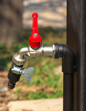 Red-handled water faucet with a hose connector, mounted on a black pipe, outdoors.