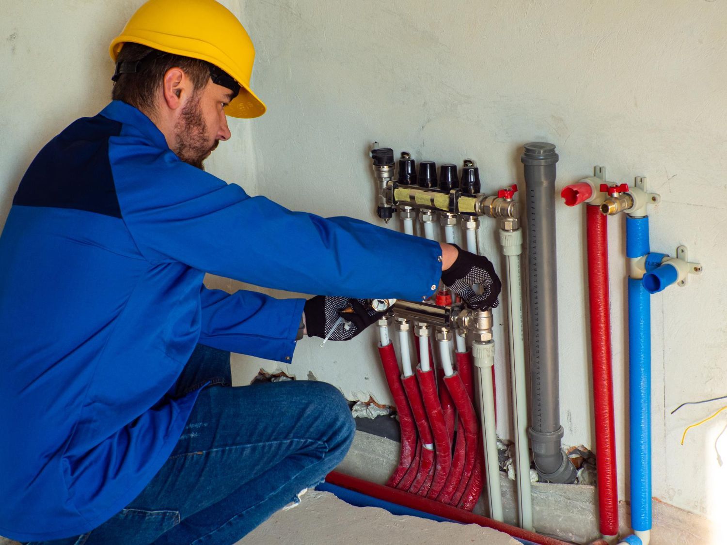 Plumber in blue work uniform, kneeling, working on pipes. Bright red and blue pipes visible.