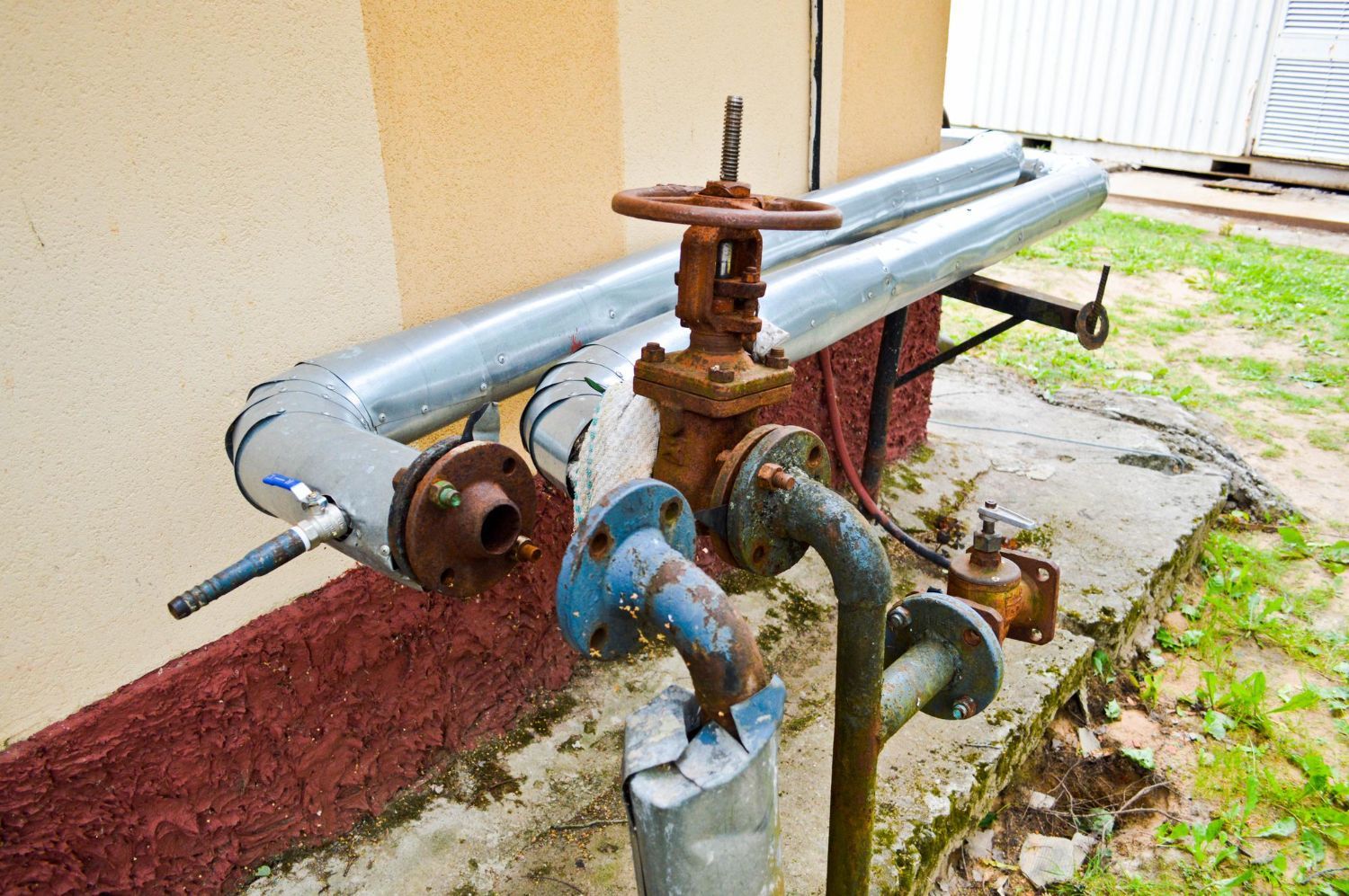 Rusty pipes with a valve, connected to a building's exterior, sitting on concrete, with some grass visible.
