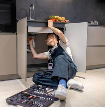 Plumber working under a kitchen sink, tools at his side. He's wearing overalls and sneakers.