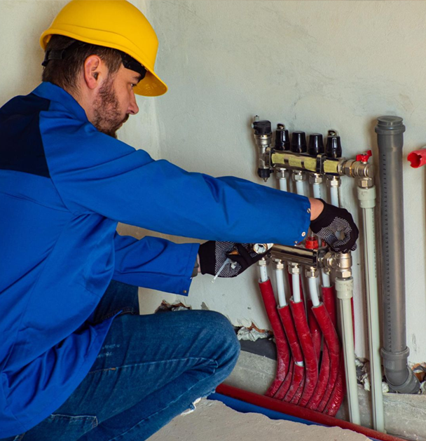 A plumber in blue workwear and a yellow hard hat works on red and white pipes.