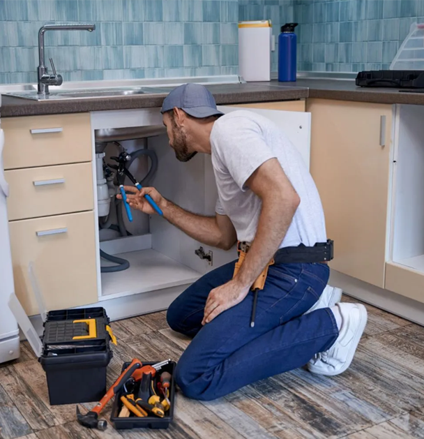 Plumber kneeling under kitchen sink, working on pipes with tools.