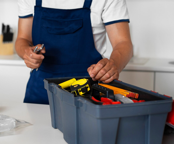 Person in blue overalls reaching into toolbox in a kitchen, holding a wrench.