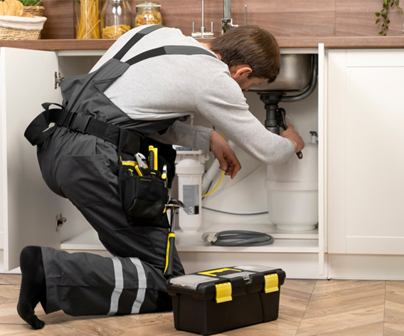 Plumber kneeling under kitchen sink, working on pipes. Black toolbox, white cabinets, tools in pouch.
