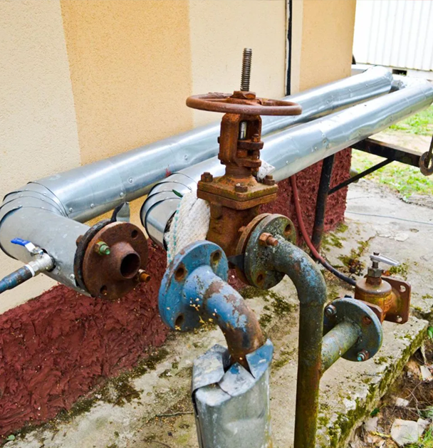 Pipes and rusty valves attached to a building's exterior wall. Blue and gray metal, insulation present.