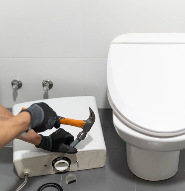 Person hammering a toilet tank in a bathroom, near a white toilet.