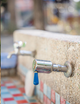 Close-up of a chrome water tap with a blue handle on a stone wall, tiled base, outdoors.