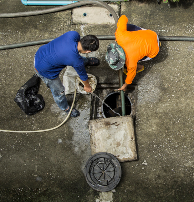 Two people working on a sewer access point; one in blue, one in orange; gray concrete, black pipes.