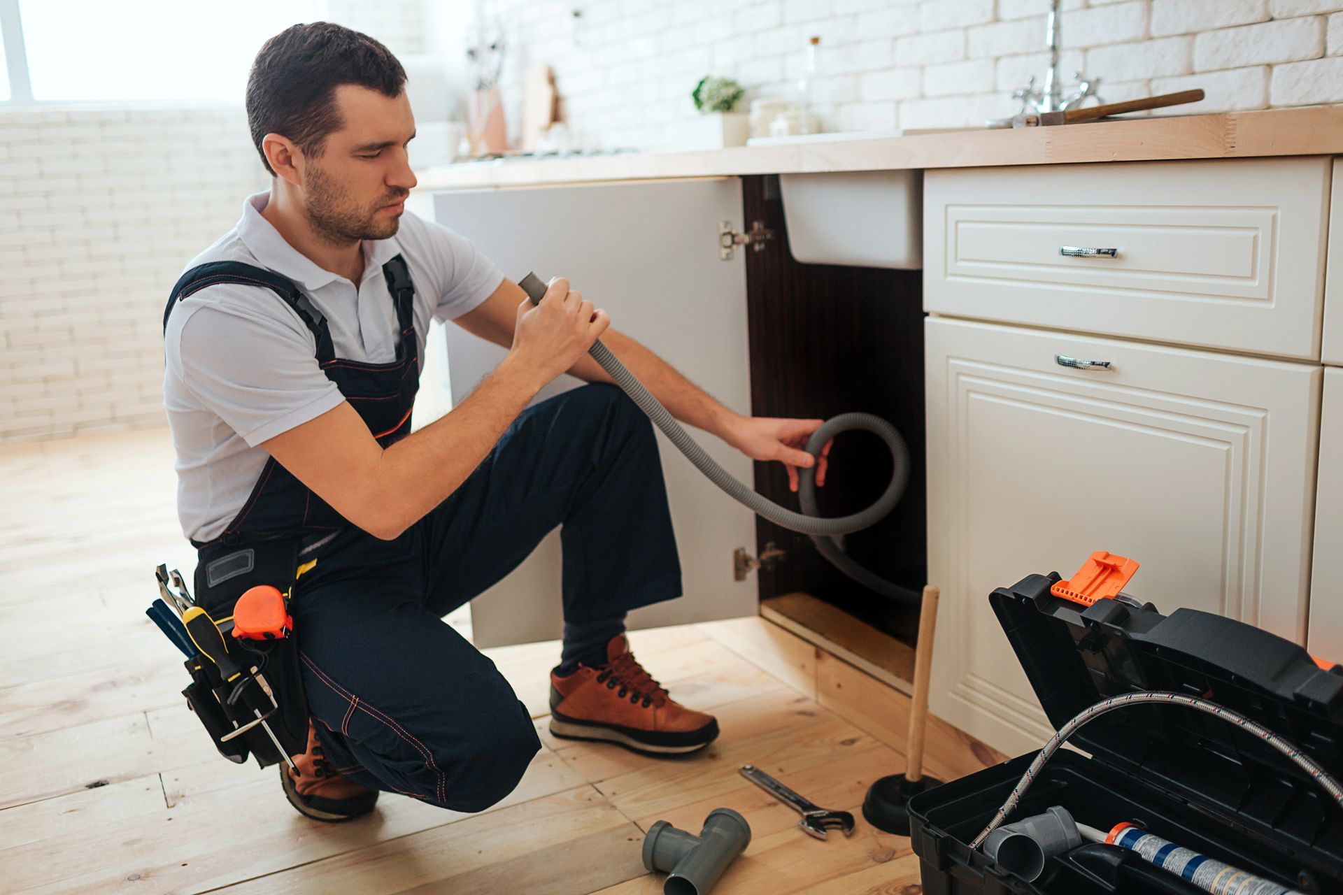 Plumber kneels under a kitchen sink, holding a pipe, with a toolbox nearby.