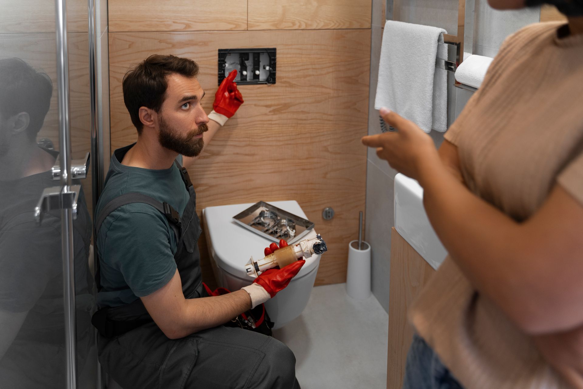 Man in red gloves fixing toilet, talking to a person in a bathroom.