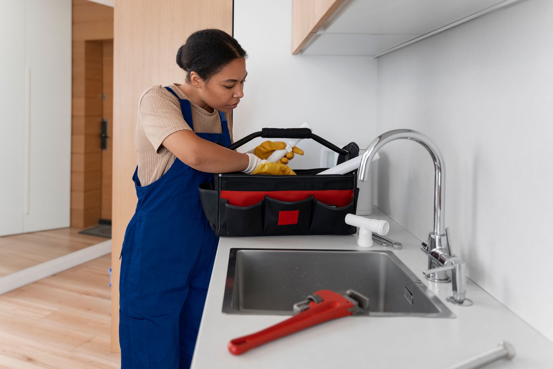 Person in blue overalls reaching into a toolbox on a kitchen counter near a sink.
