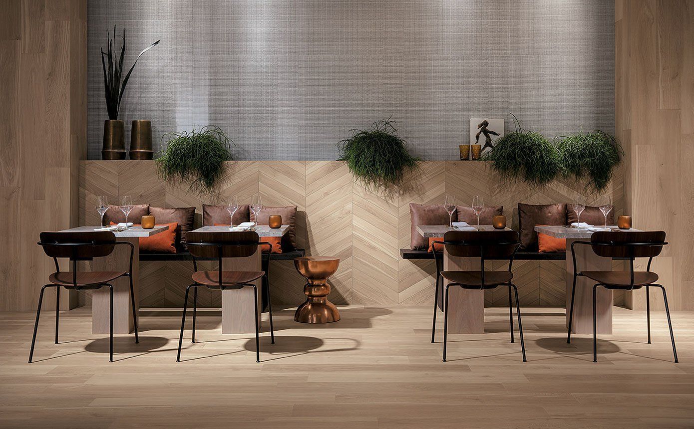 Restaurant seating area with tables, chairs, and cushioned benches, natural light. Walls are neutral, and plants provide color.