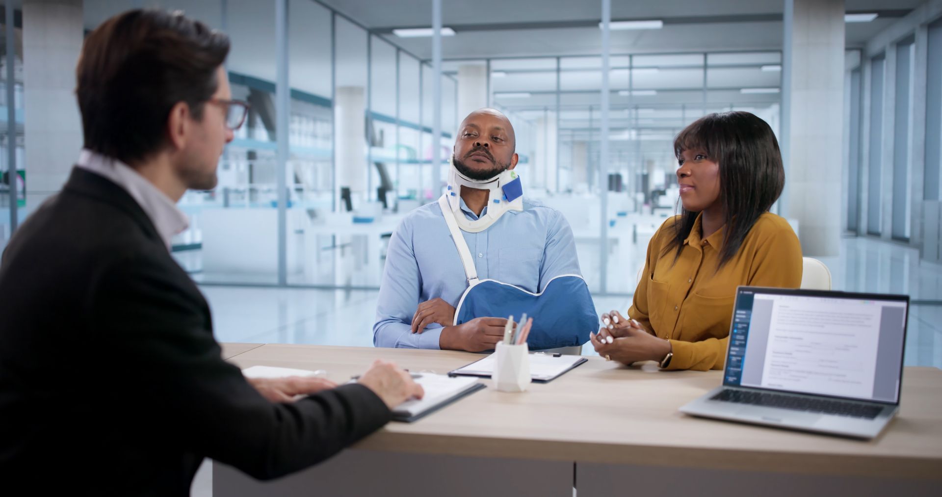 Attorney taking notes across desk from injured client with neck brace, arm sling, and companion.