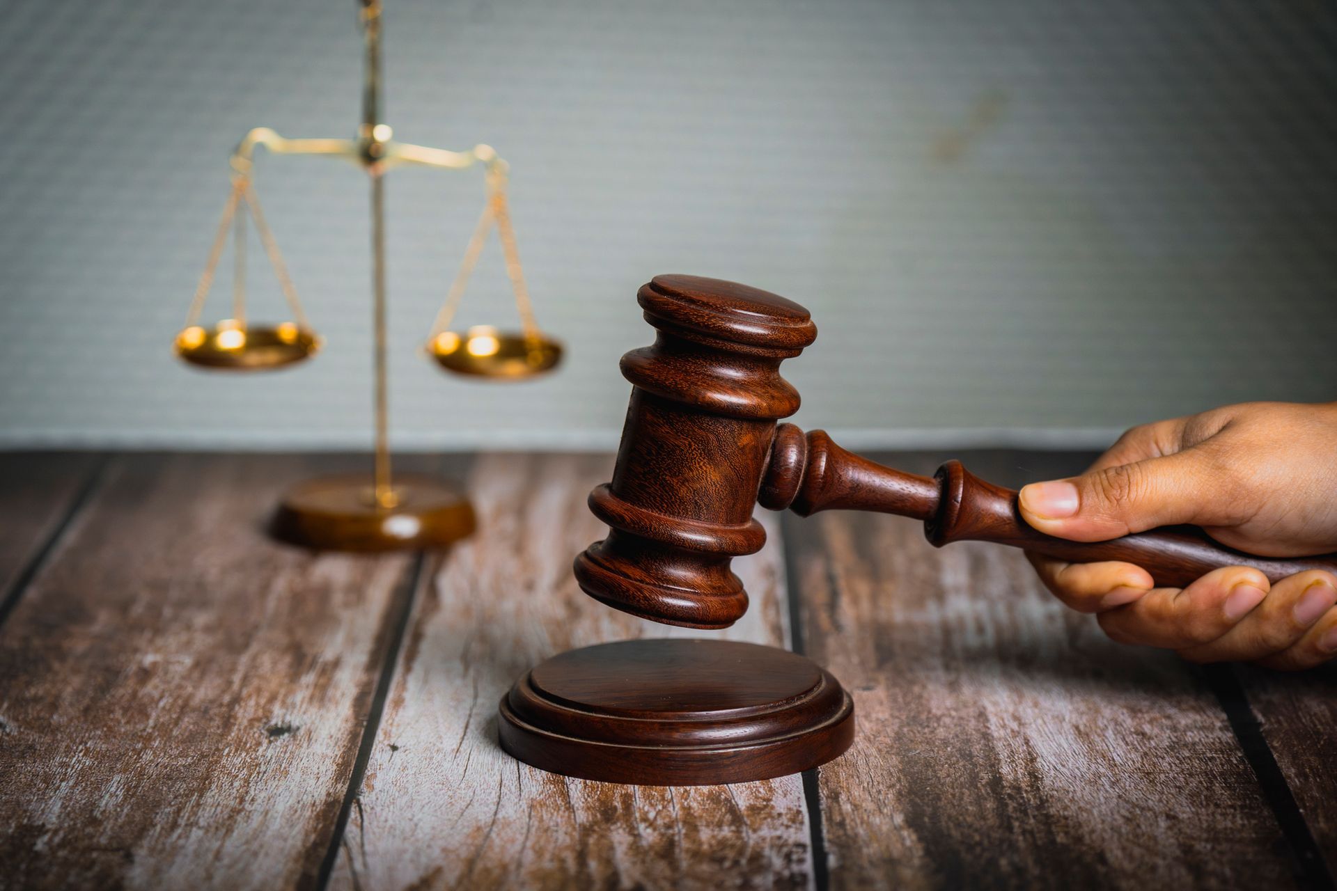 A close-up of a lawyer’s hand holding a wooden gavel with the Scales of Justice in the background.