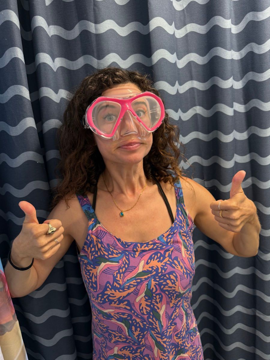 Woman in pink scuba mask and tank top gives a thumbs up in front of a wavy curtain.