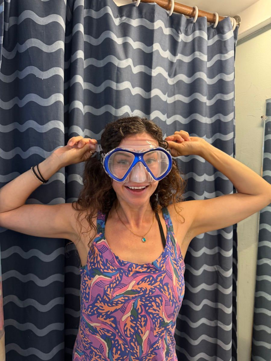 Woman wearing blue scuba mask, smiles playfully, arms up, stands in front of wavy blue curtains.