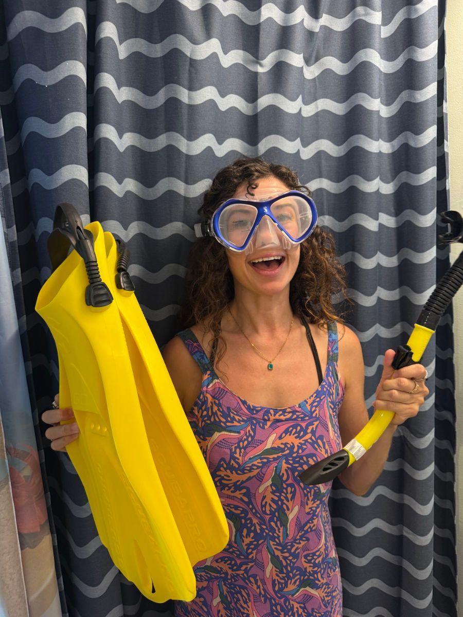 Woman in diving gear, smiling, holds yellow fins and a snorkel against a wavy curtain backdrop.