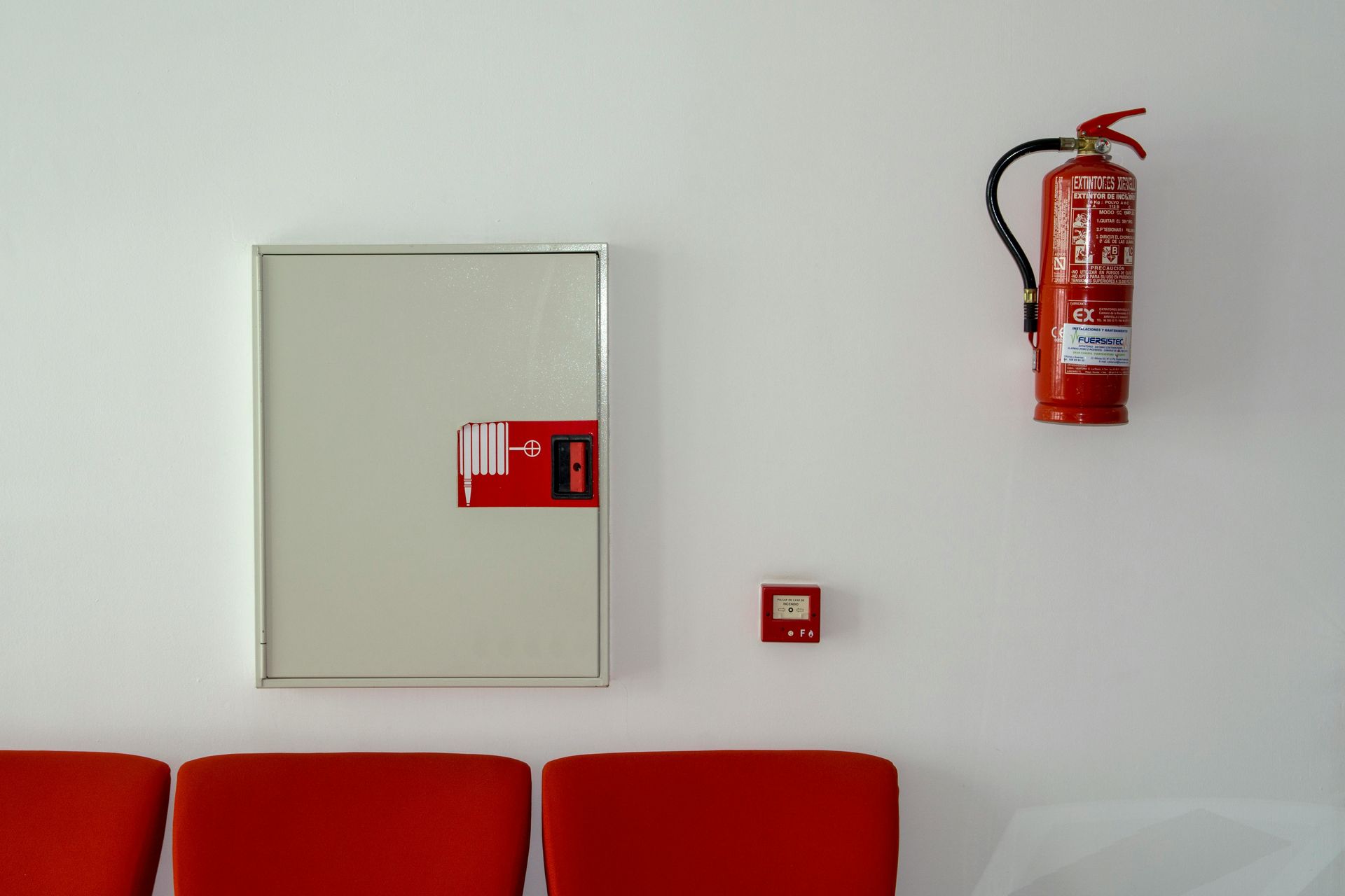 Red fire extinguisher and cabinet on white wall, with a small red alarm.