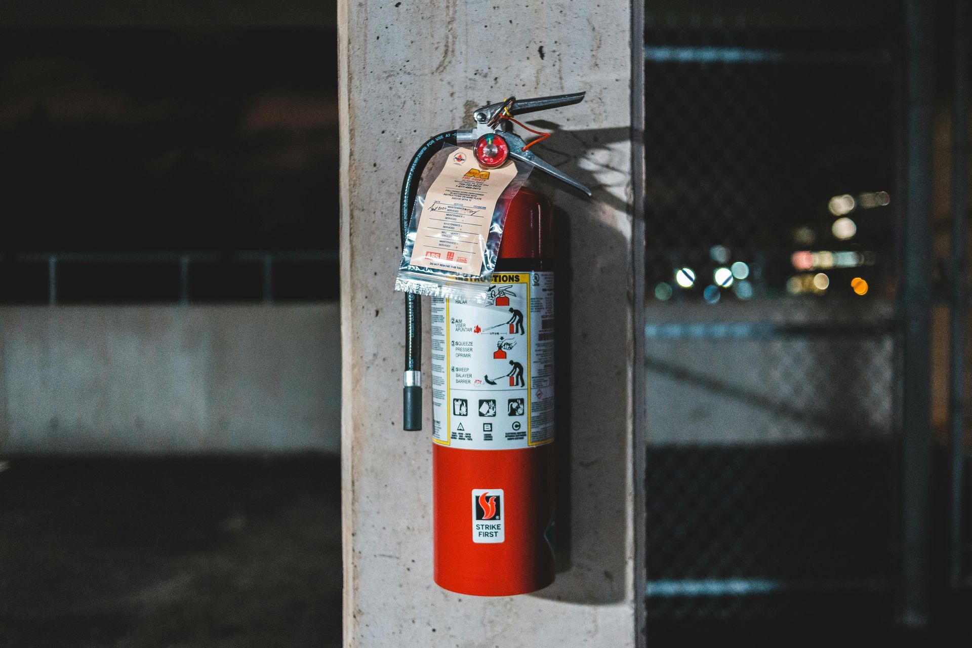 Red fire extinguisher on a concrete pillar; instructions and gauge visible. Dark background with city lights.
