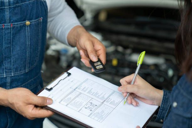 Mechanic handing a car key to a person signing paperwork in front of a car engine.
