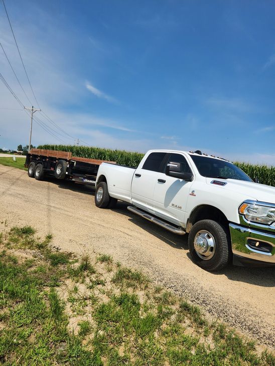 White pickup truck towing a flatbed trailer on a dirt road next to a cornfield, under a blue sky.