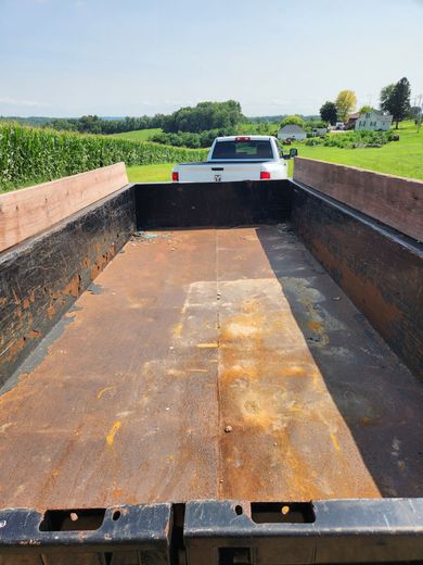 Empty, rusty black truck bed with wooden side rails; white truck in the distance, green fields and blue sky.